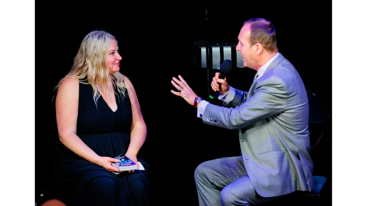 Christopher Carter holding a microphone and gesturing while facing a seated audience volunteer onstage.
